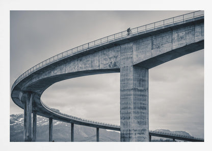 A black and white photograph of a lone cyclist riding across the top level of a massive, curving concrete bridge, which sweeps across the frame. In the background, a lower level of the bridge and distant mountains are visible under a cloudy sky. Decor