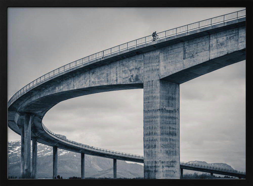 A black and white photograph of a lone cyclist riding across the top level of a massive, curving concrete bridge, which sweeps across the frame. In the background, a lower level of the bridge and distant mountains are visible under a cloudy sky. Decor