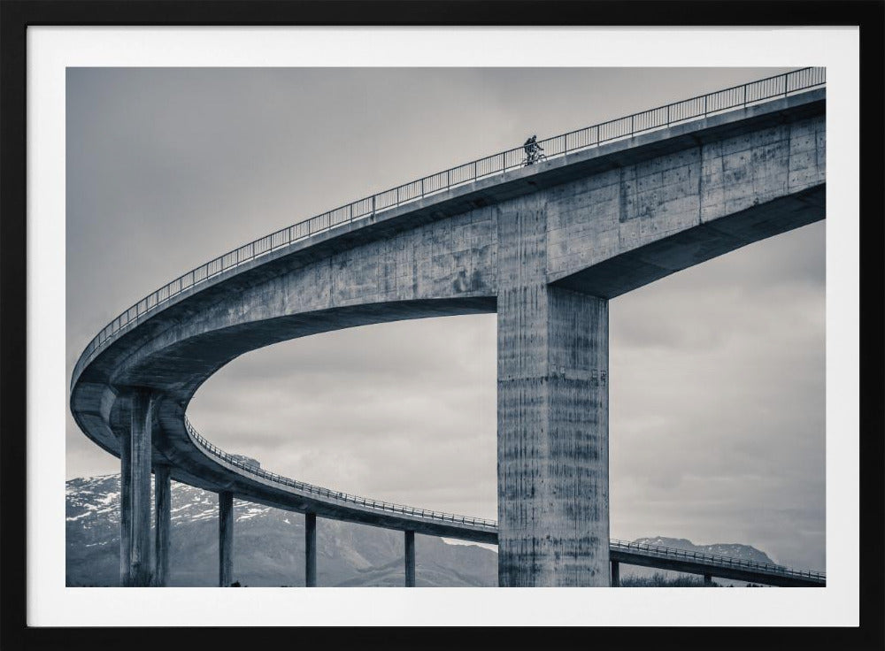 A black and white photograph of a lone cyclist riding across the top level of a massive, curving concrete bridge, which sweeps across the frame. In the background, a lower level of the bridge and distant mountains are visible under a cloudy sky. Decor