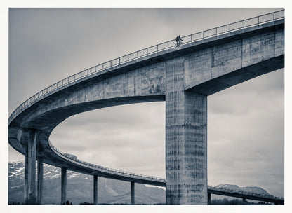 A black and white photograph of a lone cyclist riding across the top level of a massive, curving concrete bridge, which sweeps across the frame. In the background, a lower level of the bridge and distant mountains are visible under a cloudy sky. Decor