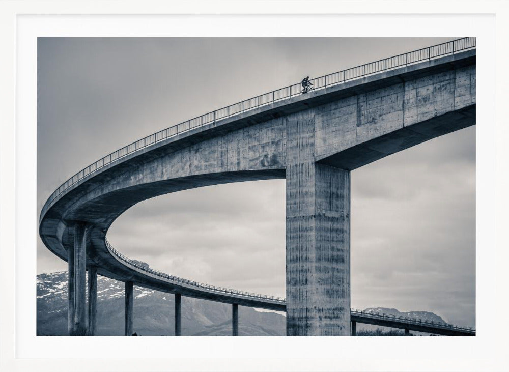 A black and white photograph of a lone cyclist riding across the top level of a massive, curving concrete bridge, which sweeps across the frame. In the background, a lower level of the bridge and distant mountains are visible under a cloudy sky. Decor