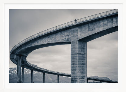 A black and white photograph of a lone cyclist riding across the top level of a massive, curving concrete bridge, which sweeps across the frame. In the background, a lower level of the bridge and distant mountains are visible under a cloudy sky. Decor