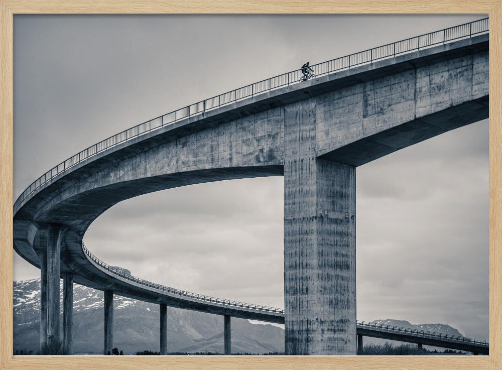 A black and white photograph of a lone cyclist riding across the top level of a massive, curving concrete bridge, which sweeps across the frame. In the background, a lower level of the bridge and distant mountains are visible under a cloudy sky. Decor