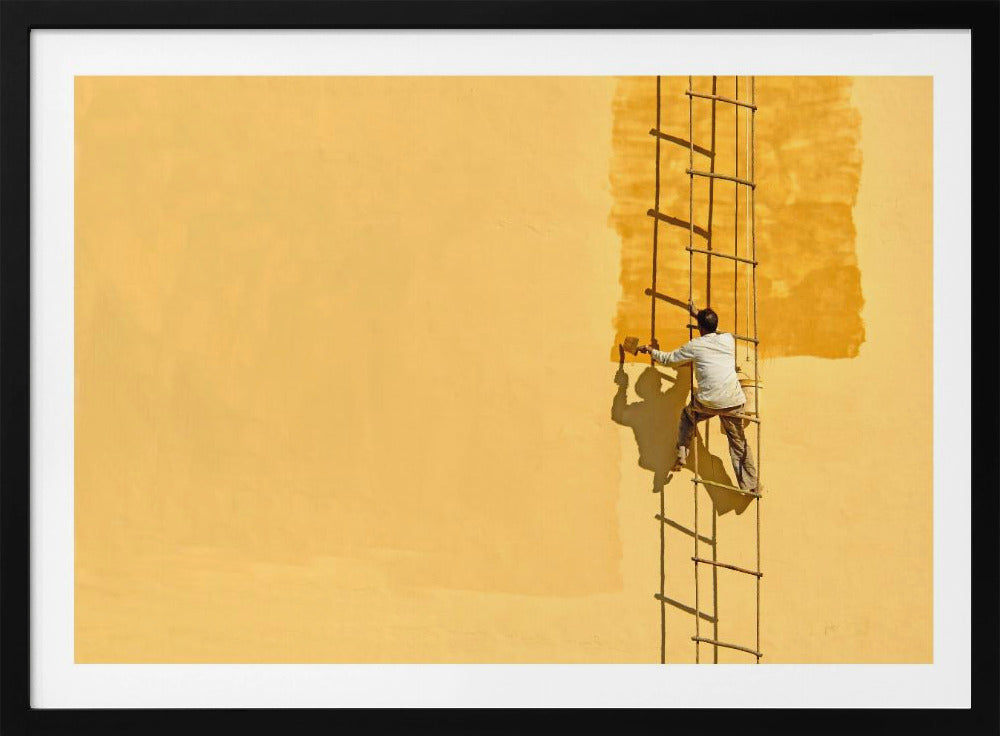 A man viewed from behind stands on a rope ladder, painting a large, bright yellow wall a slightly darker shade of yellow, casting a stark shadow in the bright sun. The image is presented within a silver frame. Decor