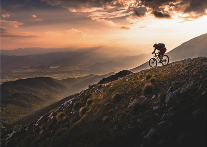 A lone mountain biker is silhouetted against a breathtaking sunset as they navigate a steep, rocky ridge. Below them, layers of mountains and a vast valley stretch into the distance under a dramatic, cloudy orange sky. Artwork