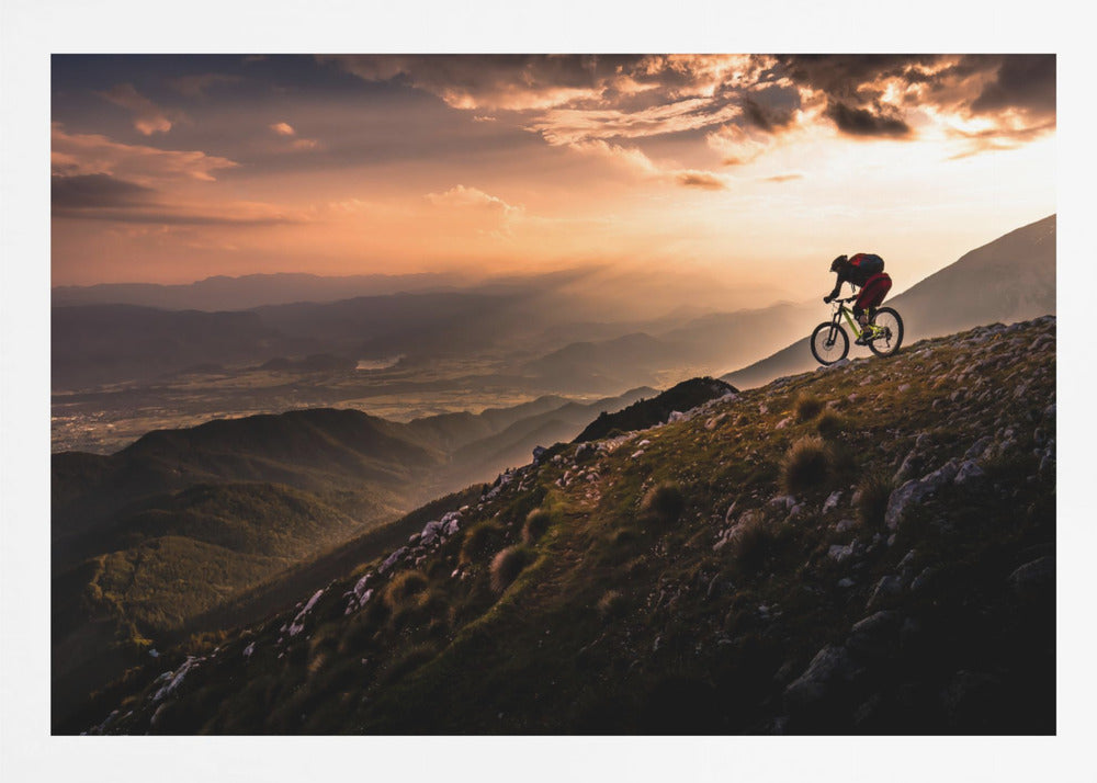 A lone mountain biker is silhouetted against a breathtaking sunset as they navigate a steep, rocky ridge. Below them, layers of mountains and a vast valley stretch into the distance under a dramatic, cloudy orange sky. Artwork