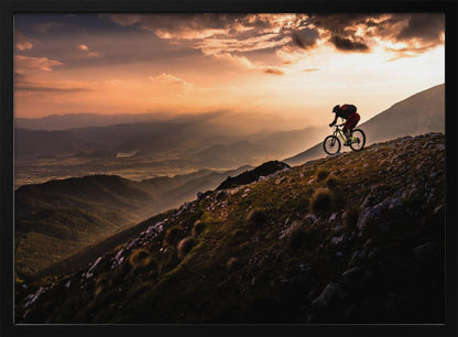 A lone mountain biker is silhouetted against a breathtaking sunset as they navigate a steep, rocky ridge. Below them, layers of mountains and a vast valley stretch into the distance under a dramatic, cloudy orange sky. Artwork