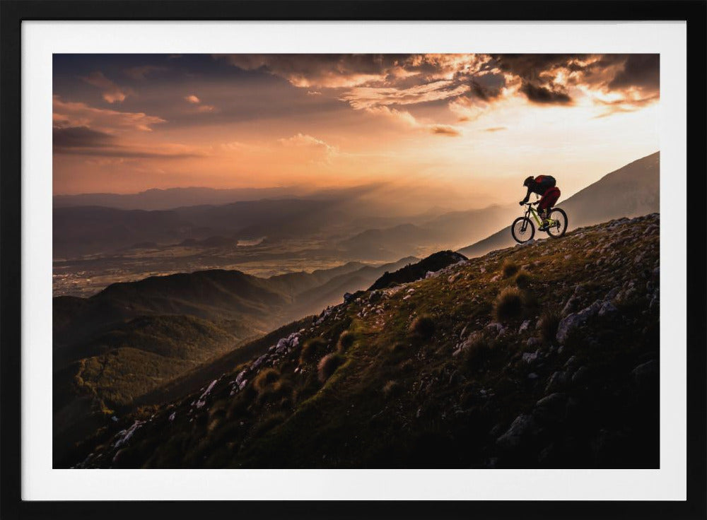 A lone mountain biker is silhouetted against a breathtaking sunset as they navigate a steep, rocky ridge. Below them, layers of mountains and a vast valley stretch into the distance under a dramatic, cloudy orange sky. Artwork