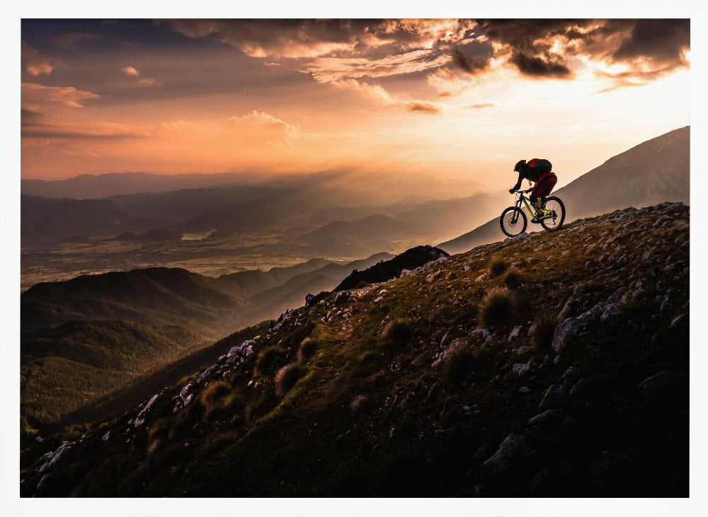 A lone mountain biker is silhouetted against a breathtaking sunset as they navigate a steep, rocky ridge. Below them, layers of mountains and a vast valley stretch into the distance under a dramatic, cloudy orange sky. Artwork