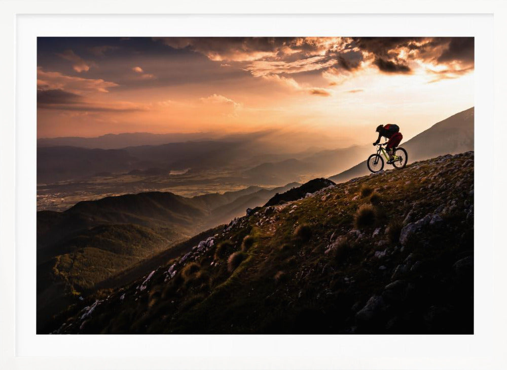 A lone mountain biker is silhouetted against a breathtaking sunset as they navigate a steep, rocky ridge. Below them, layers of mountains and a vast valley stretch into the distance under a dramatic, cloudy orange sky. Artwork