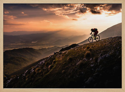 A lone mountain biker is silhouetted against a breathtaking sunset as they navigate a steep, rocky ridge. Below them, layers of mountains and a vast valley stretch into the distance under a dramatic, cloudy orange sky. Artwork