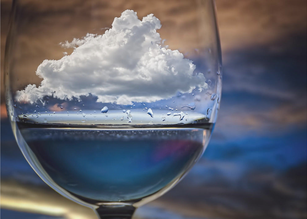 A surreal close-up of a wine glass holding blue liquid and a single white cloud, with water droplets on the glass, set against a blurred background and enclosed in a silver frame. Print