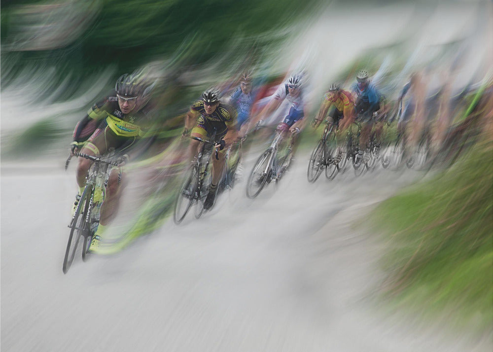 A dynamic, motion-blurred photograph of a group of cyclists in a race, captured with a slow shutter speed to create streaks of color and a sense of high speed. The lead cyclist in a yellow and black jersey is the most defined figure, leaning into a turn on a grey road flanked by blurry green foliage. The entire image is enclosed in a silver frame. Decor