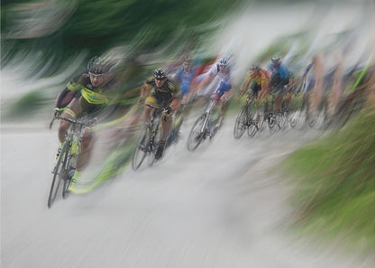 A dynamic, motion-blurred photograph of a group of cyclists in a race, captured with a slow shutter speed to create streaks of color and a sense of high speed. The lead cyclist in a yellow and black jersey is the most defined figure, leaning into a turn on a grey road flanked by blurry green foliage. The entire image is enclosed in a silver frame. Decor