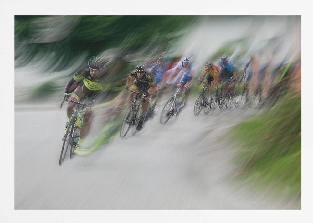 A dynamic, motion-blurred photograph of a group of cyclists in a race, captured with a slow shutter speed to create streaks of color and a sense of high speed. The lead cyclist in a yellow and black jersey is the most defined figure, leaning into a turn on a grey road flanked by blurry green foliage. The entire image is enclosed in a silver frame. Decor