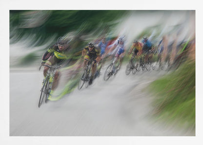 A dynamic, motion-blurred photograph of a group of cyclists in a race, captured with a slow shutter speed to create streaks of color and a sense of high speed. The lead cyclist in a yellow and black jersey is the most defined figure, leaning into a turn on a grey road flanked by blurry green foliage. The entire image is enclosed in a silver frame. Decor