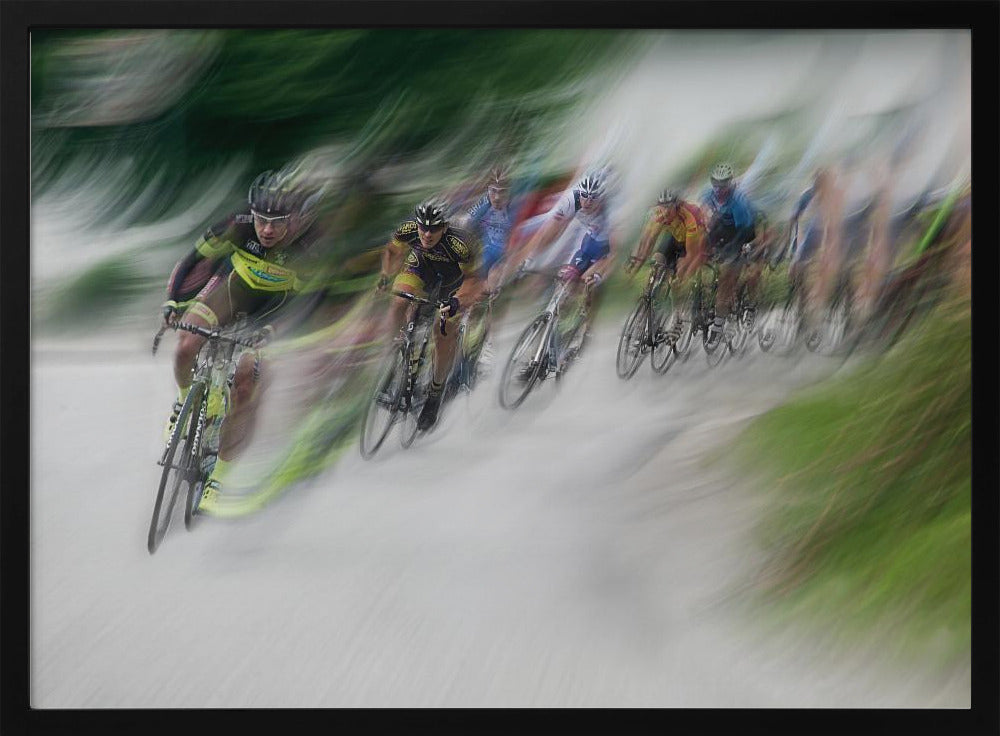 A dynamic, motion-blurred photograph of a group of cyclists in a race, captured with a slow shutter speed to create streaks of color and a sense of high speed. The lead cyclist in a yellow and black jersey is the most defined figure, leaning into a turn on a grey road flanked by blurry green foliage. The entire image is enclosed in a silver frame. Decor