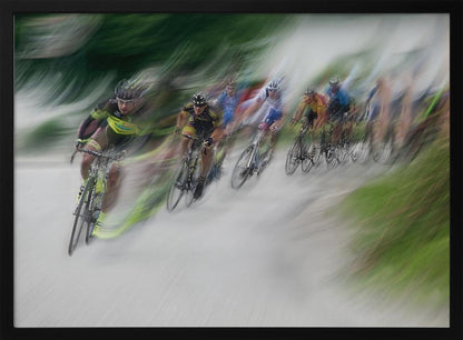 A dynamic, motion-blurred photograph of a group of cyclists in a race, captured with a slow shutter speed to create streaks of color and a sense of high speed. The lead cyclist in a yellow and black jersey is the most defined figure, leaning into a turn on a grey road flanked by blurry green foliage. The entire image is enclosed in a silver frame. Decor