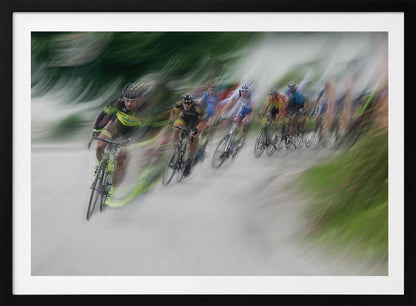 A dynamic, motion-blurred photograph of a group of cyclists in a race, captured with a slow shutter speed to create streaks of color and a sense of high speed. The lead cyclist in a yellow and black jersey is the most defined figure, leaning into a turn on a grey road flanked by blurry green foliage. The entire image is enclosed in a silver frame. Decor