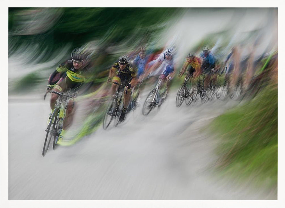 A dynamic, motion-blurred photograph of a group of cyclists in a race, captured with a slow shutter speed to create streaks of color and a sense of high speed. The lead cyclist in a yellow and black jersey is the most defined figure, leaning into a turn on a grey road flanked by blurry green foliage. The entire image is enclosed in a silver frame. Decor
