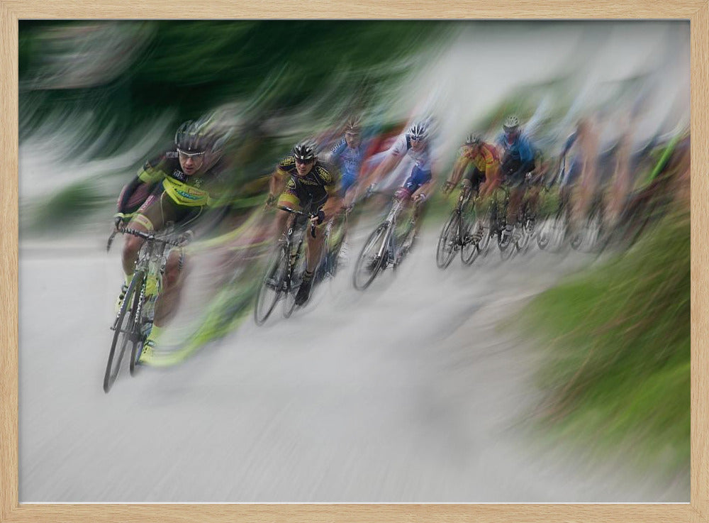 A dynamic, motion-blurred photograph of a group of cyclists in a race, captured with a slow shutter speed to create streaks of color and a sense of high speed. The lead cyclist in a yellow and black jersey is the most defined figure, leaning into a turn on a grey road flanked by blurry green foliage. The entire image is enclosed in a silver frame. Decor