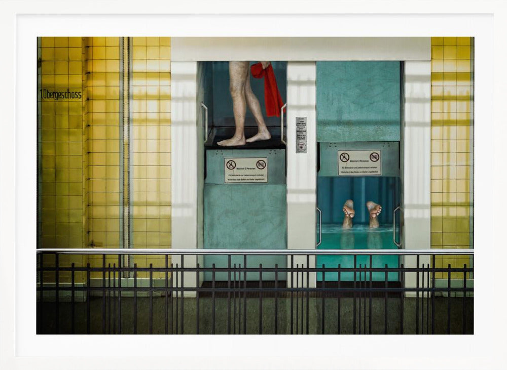 A surreal, framed artwork depicting two teal-colored vertical cabins against a yellow tiled wall. In the left cabin, a person's bare legs are visible as they hold a red towel. In the right cabin, which is filled with water, only a pair of feet can be seen submerged. A black railing is in the foreground, adding to the strange, layered composition. Artwork