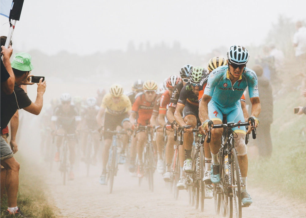 A framed photograph of a peloton of professional cyclists racing down a dusty dirt road. The lead cyclist is in sharp focus, wearing a bright turquoise uniform and a helmet, with other racers in yellow and red jerseys blurred behind him. A spectator on the left is taking a photo with a smartphone. Poster