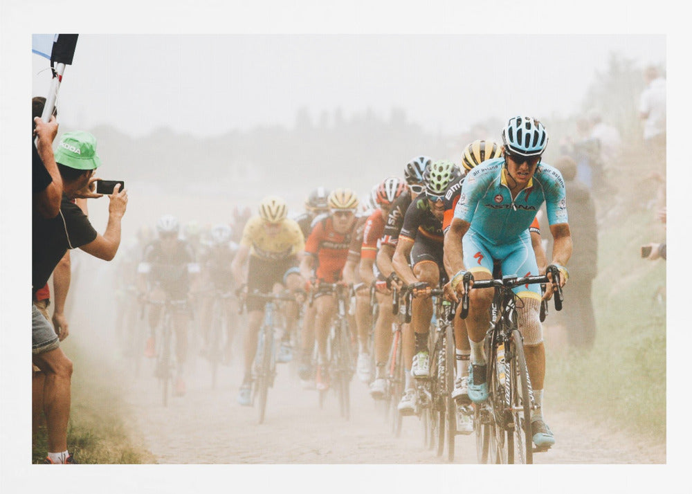 A framed photograph of a peloton of professional cyclists racing down a dusty dirt road. The lead cyclist is in sharp focus, wearing a bright turquoise uniform and a helmet, with other racers in yellow and red jerseys blurred behind him. A spectator on the left is taking a photo with a smartphone. Poster