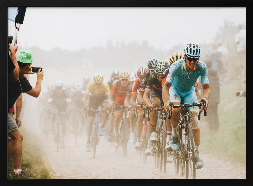 A framed photograph of a peloton of professional cyclists racing down a dusty dirt road. The lead cyclist is in sharp focus, wearing a bright turquoise uniform and a helmet, with other racers in yellow and red jerseys blurred behind him. A spectator on the left is taking a photo with a smartphone. Poster