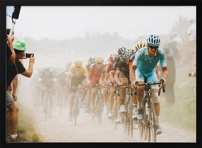 A framed photograph of a peloton of professional cyclists racing down a dusty dirt road. The lead cyclist is in sharp focus, wearing a bright turquoise uniform and a helmet, with other racers in yellow and red jerseys blurred behind him. A spectator on the left is taking a photo with a smartphone. Poster