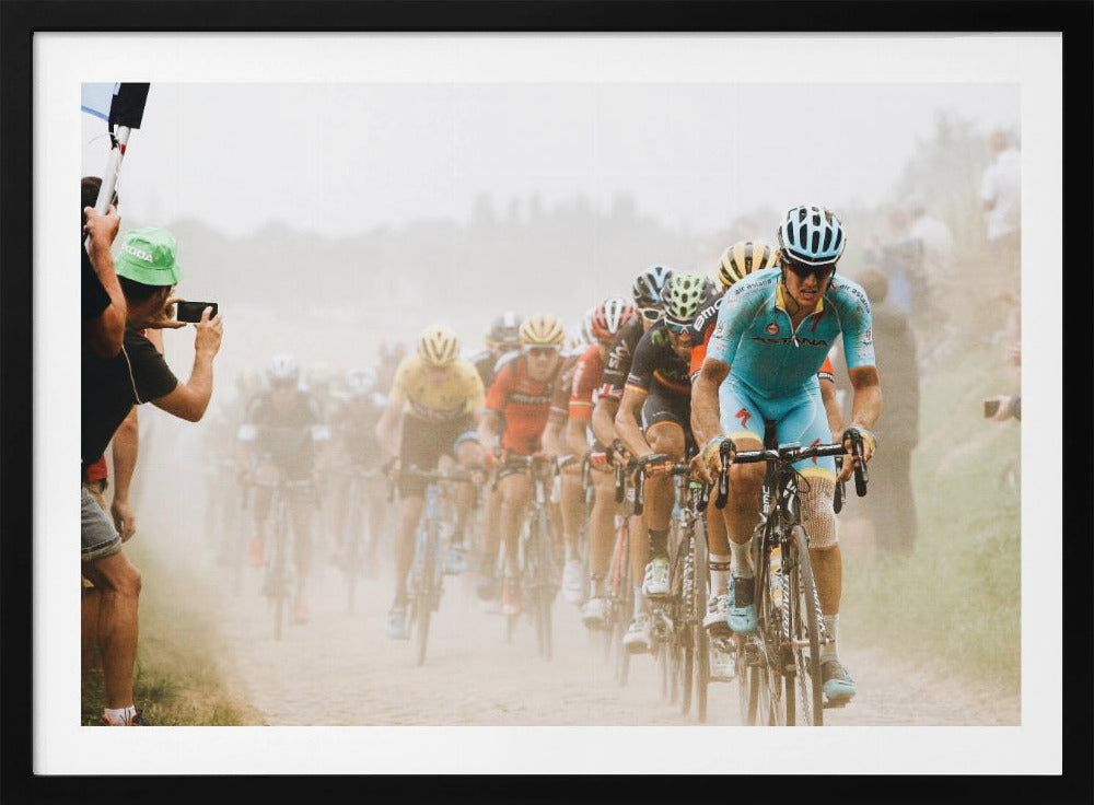 A framed photograph of a peloton of professional cyclists racing down a dusty dirt road. The lead cyclist is in sharp focus, wearing a bright turquoise uniform and a helmet, with other racers in yellow and red jerseys blurred behind him. A spectator on the left is taking a photo with a smartphone. Poster