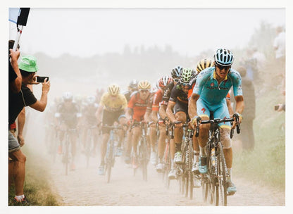 A framed photograph of a peloton of professional cyclists racing down a dusty dirt road. The lead cyclist is in sharp focus, wearing a bright turquoise uniform and a helmet, with other racers in yellow and red jerseys blurred behind him. A spectator on the left is taking a photo with a smartphone. Poster