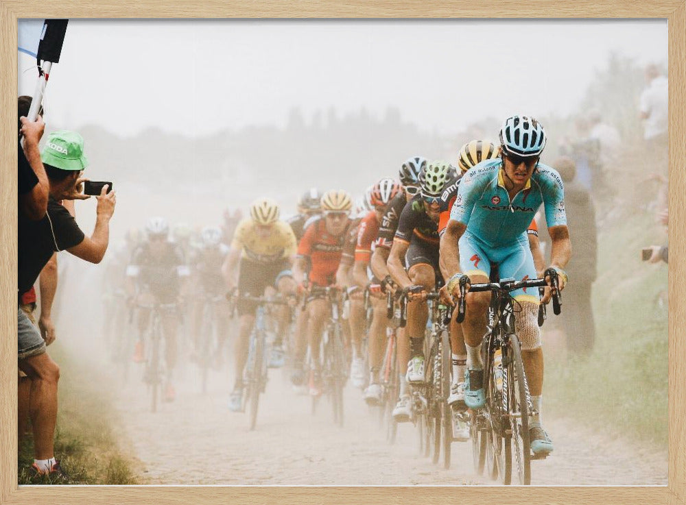 A framed photograph of a peloton of professional cyclists racing down a dusty dirt road. The lead cyclist is in sharp focus, wearing a bright turquoise uniform and a helmet, with other racers in yellow and red jerseys blurred behind him. A spectator on the left is taking a photo with a smartphone. Poster