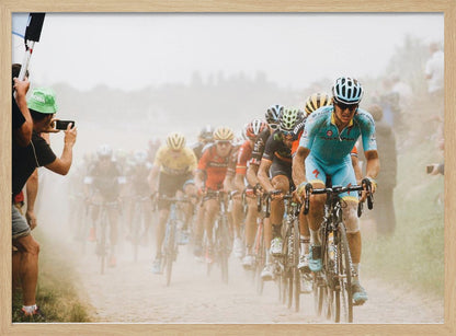 A framed photograph of a peloton of professional cyclists racing down a dusty dirt road. The lead cyclist is in sharp focus, wearing a bright turquoise uniform and a helmet, with other racers in yellow and red jerseys blurred behind him. A spectator on the left is taking a photo with a smartphone. Poster