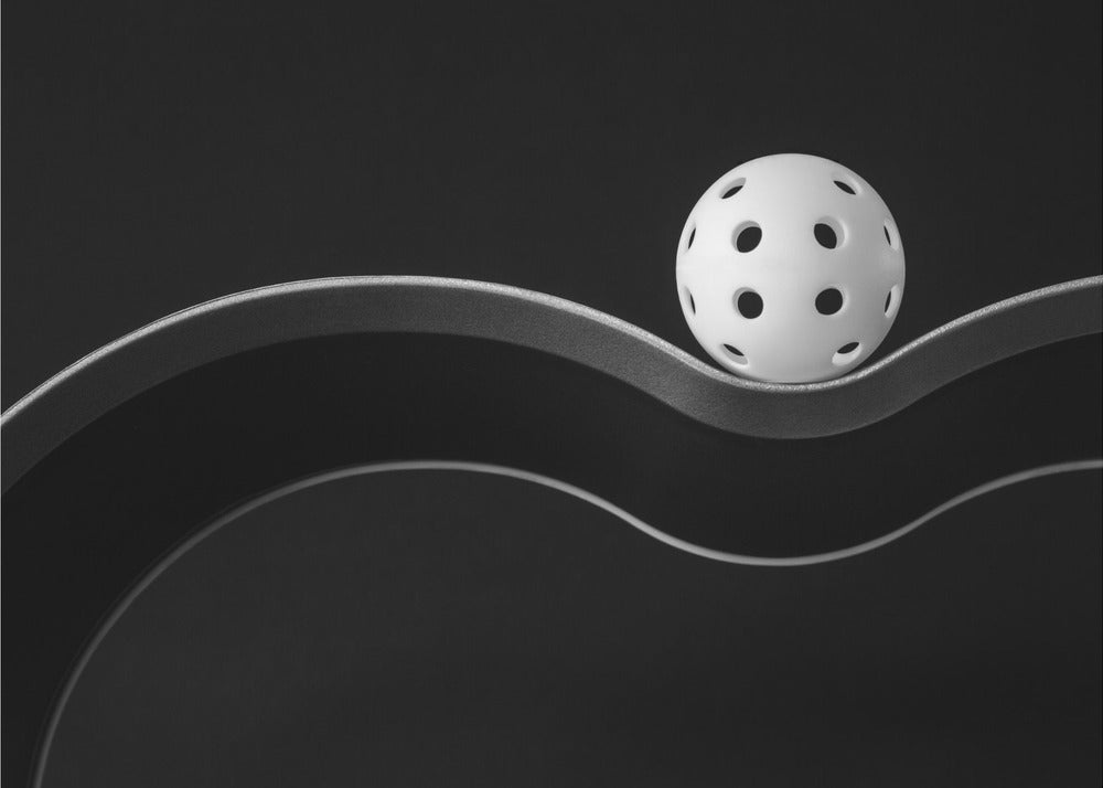 A minimalist black and white photograph of a white pickleball resting perfectly balanced on a thick, wavy, textured line against a solid black background. Below it, a thin white line mimics the curve. The entire piece is framed in silver. Poster