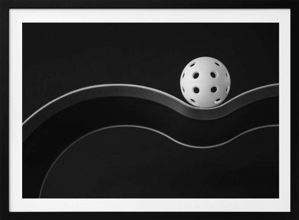 A minimalist black and white photograph of a white pickleball resting perfectly balanced on a thick, wavy, textured line against a solid black background. Below it, a thin white line mimics the curve. The entire piece is framed in silver. Poster