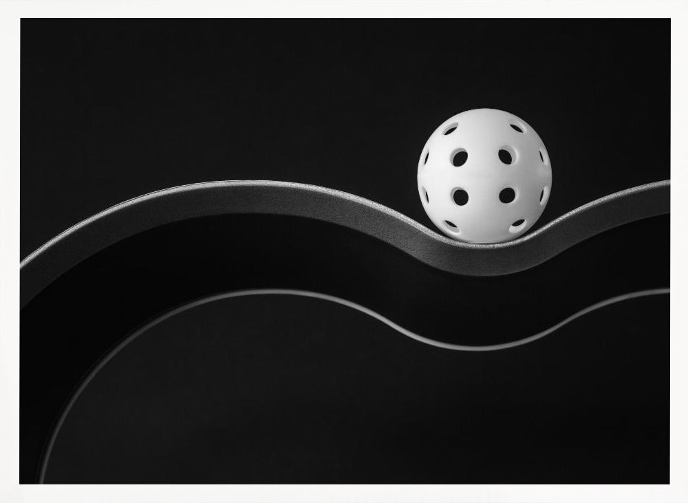 A minimalist black and white photograph of a white pickleball resting perfectly balanced on a thick, wavy, textured line against a solid black background. Below it, a thin white line mimics the curve. The entire piece is framed in silver. Poster