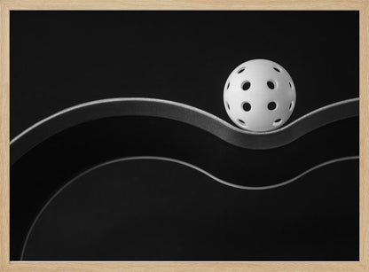 A minimalist black and white photograph of a white pickleball resting perfectly balanced on a thick, wavy, textured line against a solid black background. Below it, a thin white line mimics the curve. The entire piece is framed in silver. Poster