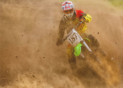 An action shot of a motocross rider in full red and yellow gear, speeding through a dirt track and kicking up a large cloud of brown dust and debris. Print