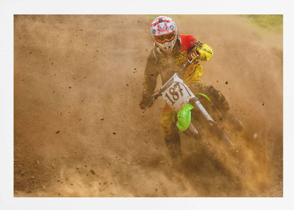 An action shot of a motocross rider in full red and yellow gear, speeding through a dirt track and kicking up a large cloud of brown dust and debris. Print