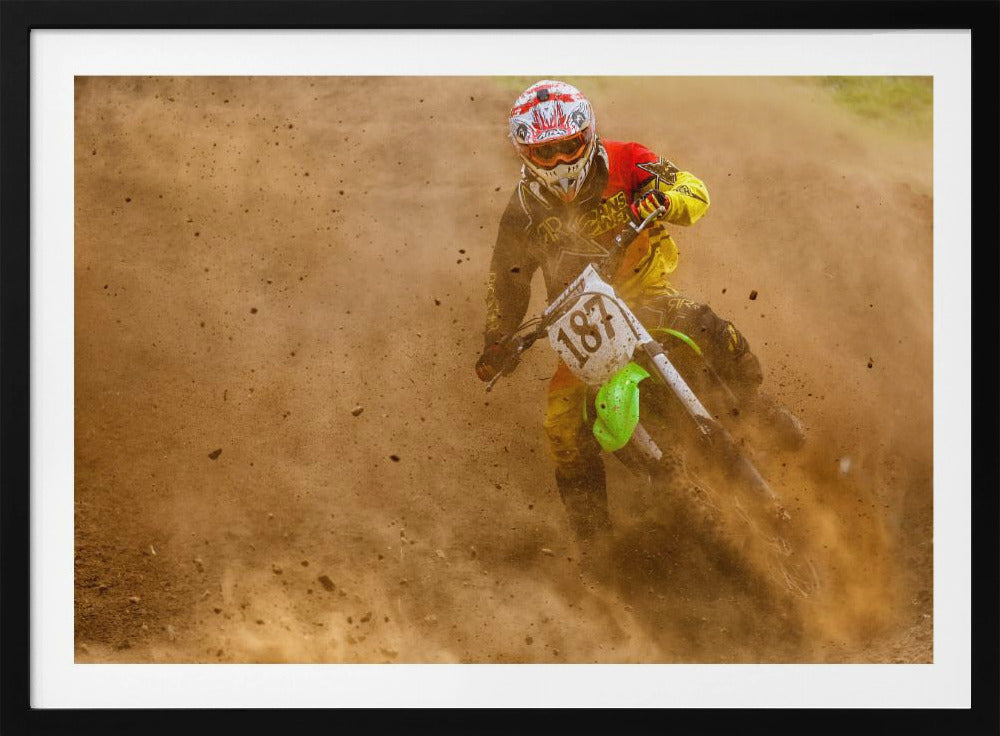 An action shot of a motocross rider in full red and yellow gear, speeding through a dirt track and kicking up a large cloud of brown dust and debris. Print