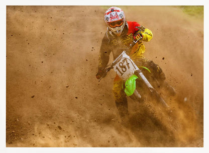 An action shot of a motocross rider in full red and yellow gear, speeding through a dirt track and kicking up a large cloud of brown dust and debris. Print