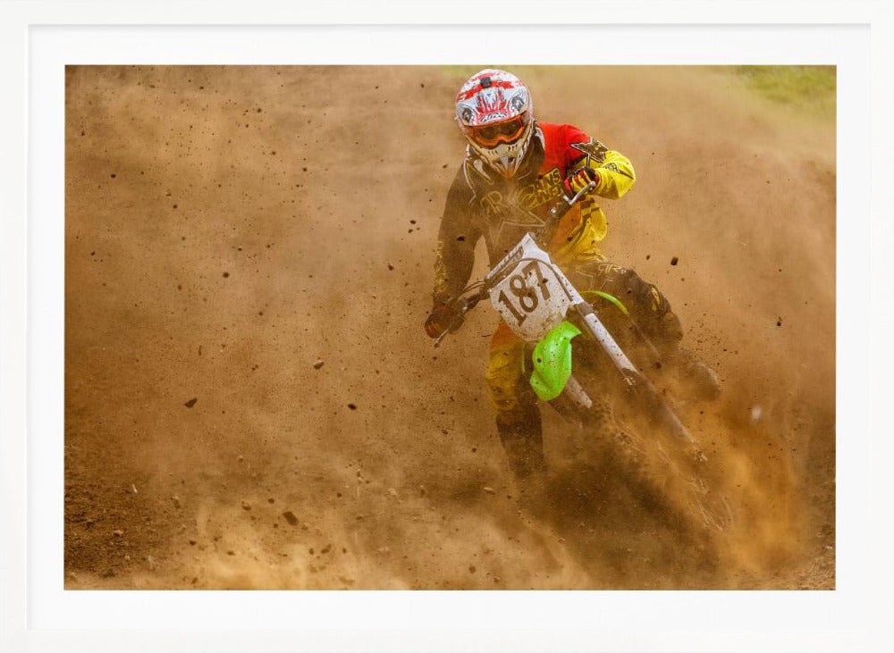 An action shot of a motocross rider in full red and yellow gear, speeding through a dirt track and kicking up a large cloud of brown dust and debris. Print
