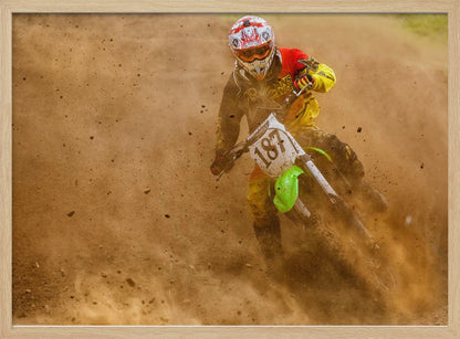 An action shot of a motocross rider in full red and yellow gear, speeding through a dirt track and kicking up a large cloud of brown dust and debris. Print
