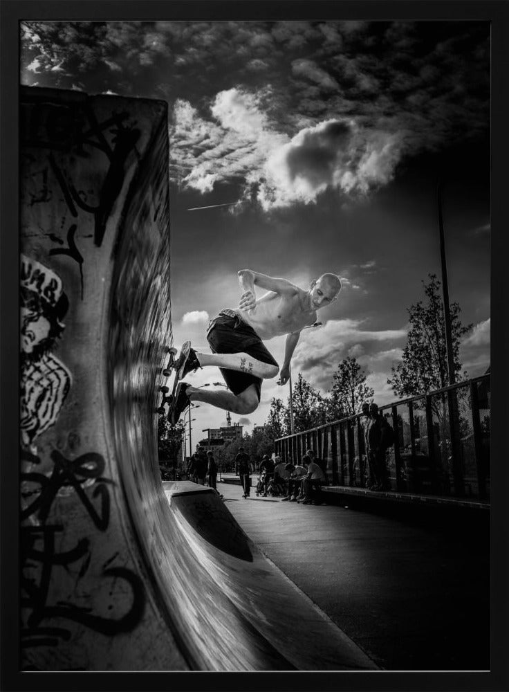 A dramatic black and white, low-angle action shot of a shirtless skateboarder suspended in mid-air at the peak of a graffiti-covered skate ramp, against a backdrop of a cloudy sky. Decor