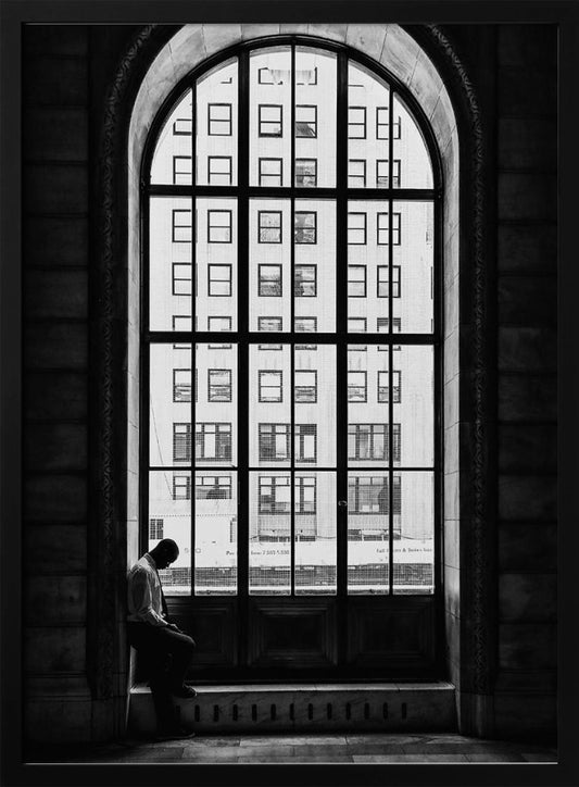 A black and white photograph of a solitary man sitting with his head bowed on the ledge of a massive arched window. The window's dark mullions create a grid over the bright view of another building outside, while the man remains in the shadows, creating a pensive and lonely mood. Poster