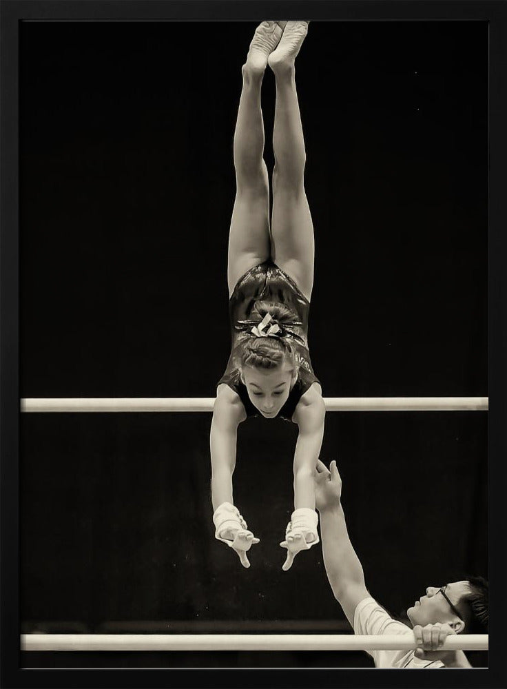 A dramatic black and white photograph of a young female gymnast performing a flawless handstand on the top of two parallel bars. Her body is perfectly vertical against a stark black background. Below, a male coach looks up at her with focused attention, his hand raised nearby, ready to spot her. Decor