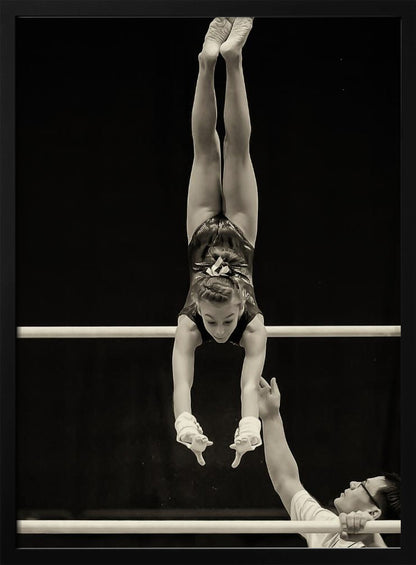 A dramatic black and white photograph of a young female gymnast performing a flawless handstand on the top of two parallel bars. Her body is perfectly vertical against a stark black background. Below, a male coach looks up at her with focused attention, his hand raised nearby, ready to spot her. Decor
