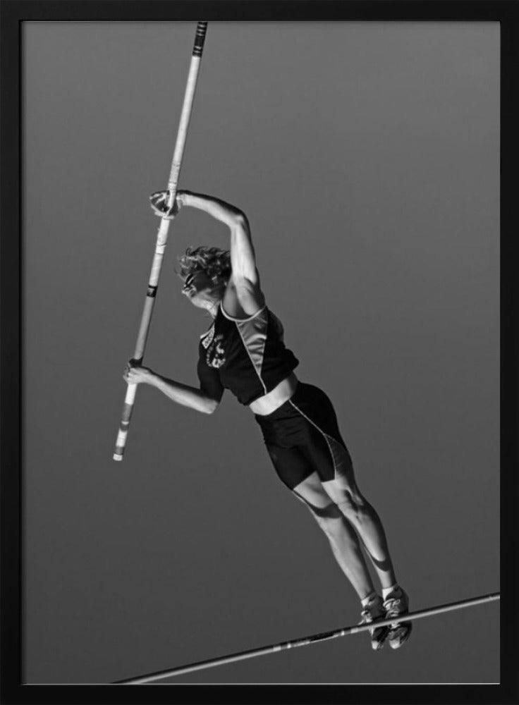 A dynamic black and white, low-angle shot of a female pole vaulter in mid-action. She is suspended in the air, gripping the pole as she prepares to clear the high bar, with her muscular form silhouetted against a plain sky. Print