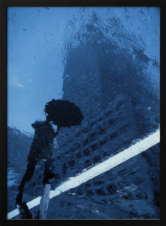 A moody, blue-toned photograph of a reflection in a rainy puddle. A black silhouette of a person holding an umbrella and a distorted brick building are visible, with a single white line crossing the wet street. Poster